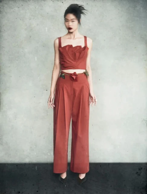 Full-length shot of a woman in an orange two-piece outfit with a floral belt posing in front of a textured grey background. Fashion editorial.