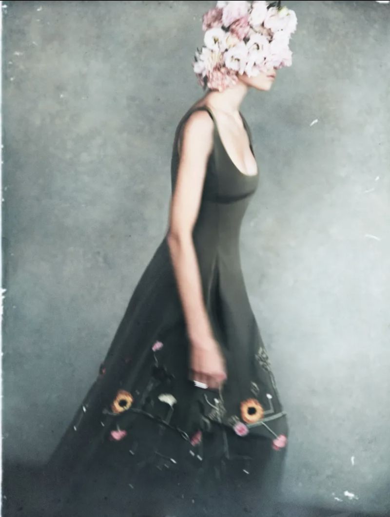 A woman in an olive dress with floral embellishments is wearing a pink and white floral headpiece in a blurred, dreamy aesthetic.