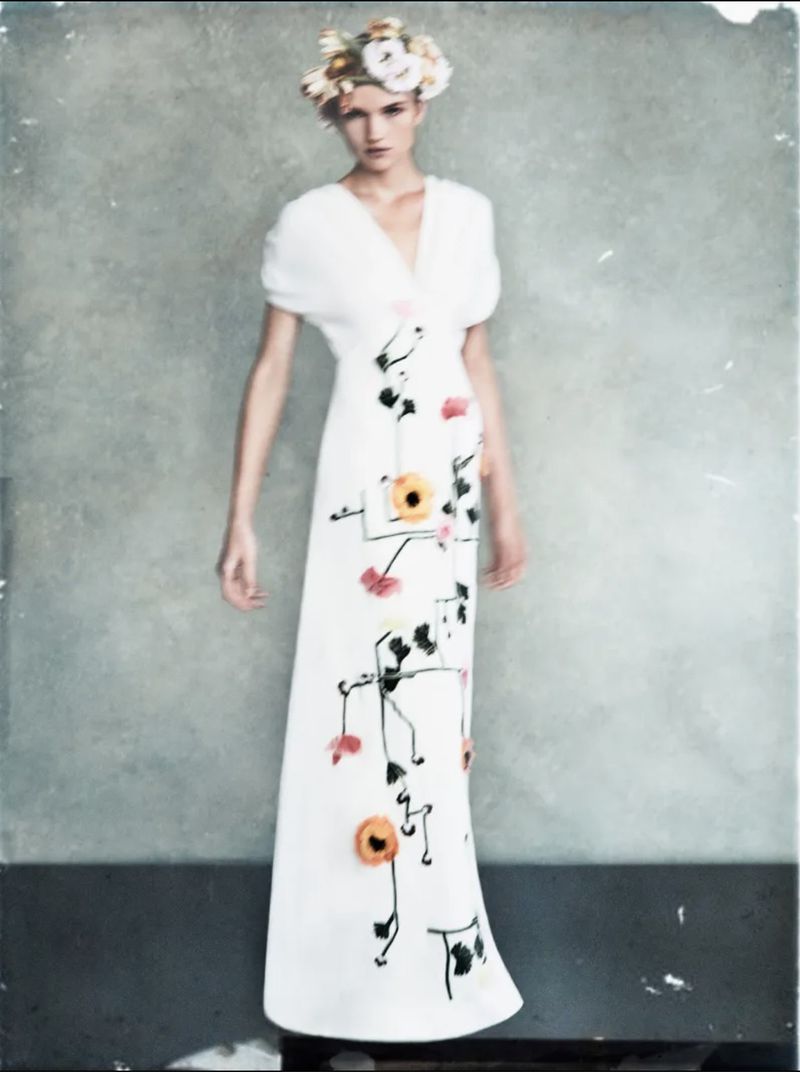 A woman in a long white dress with floral designs and a floral headpiece poses in front of a neutral background in a fashion shot.