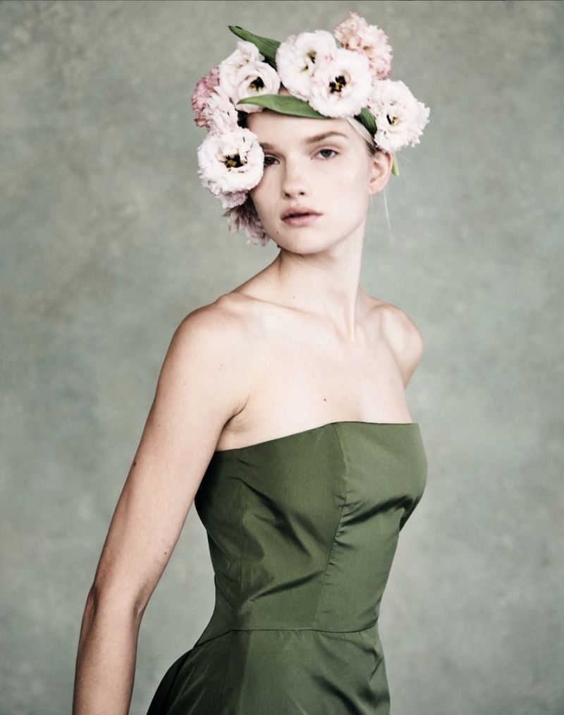 Woman wearing a green strapless dress and a floral headpiece poses in front of a mottled grey and green background in a studio portrait.