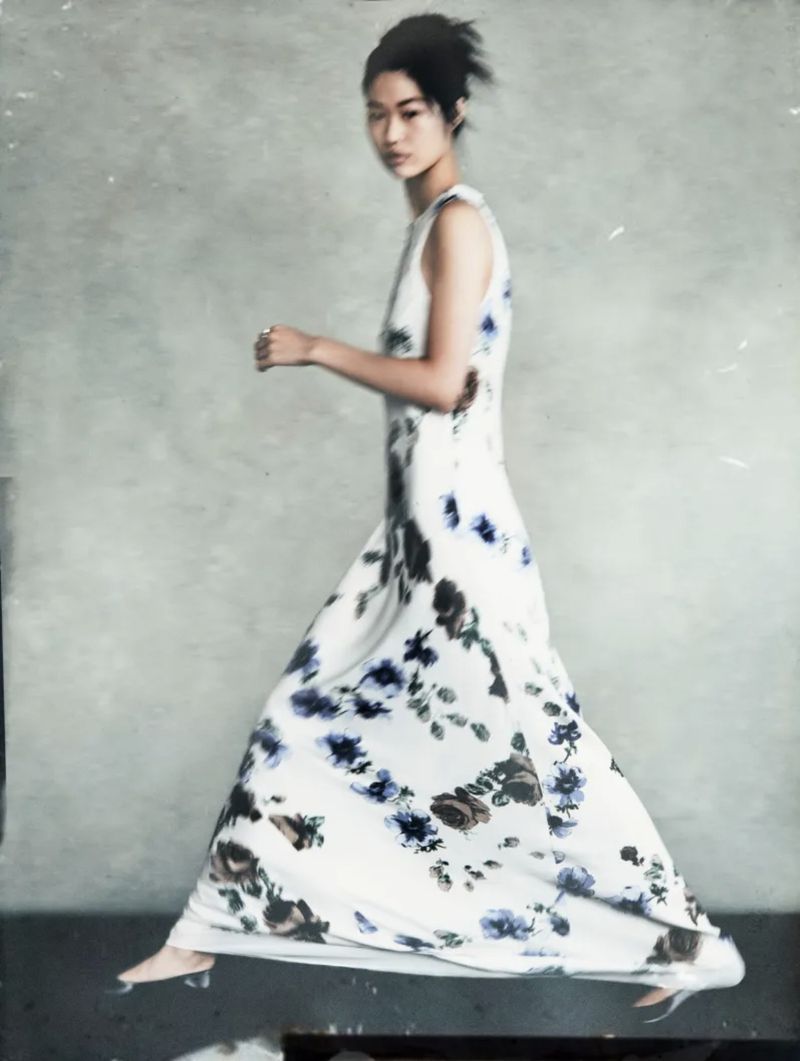 Asian woman in floral print dress mid-stride, fashion portrait. Full-length shot with natural lighting against a muted grey background.