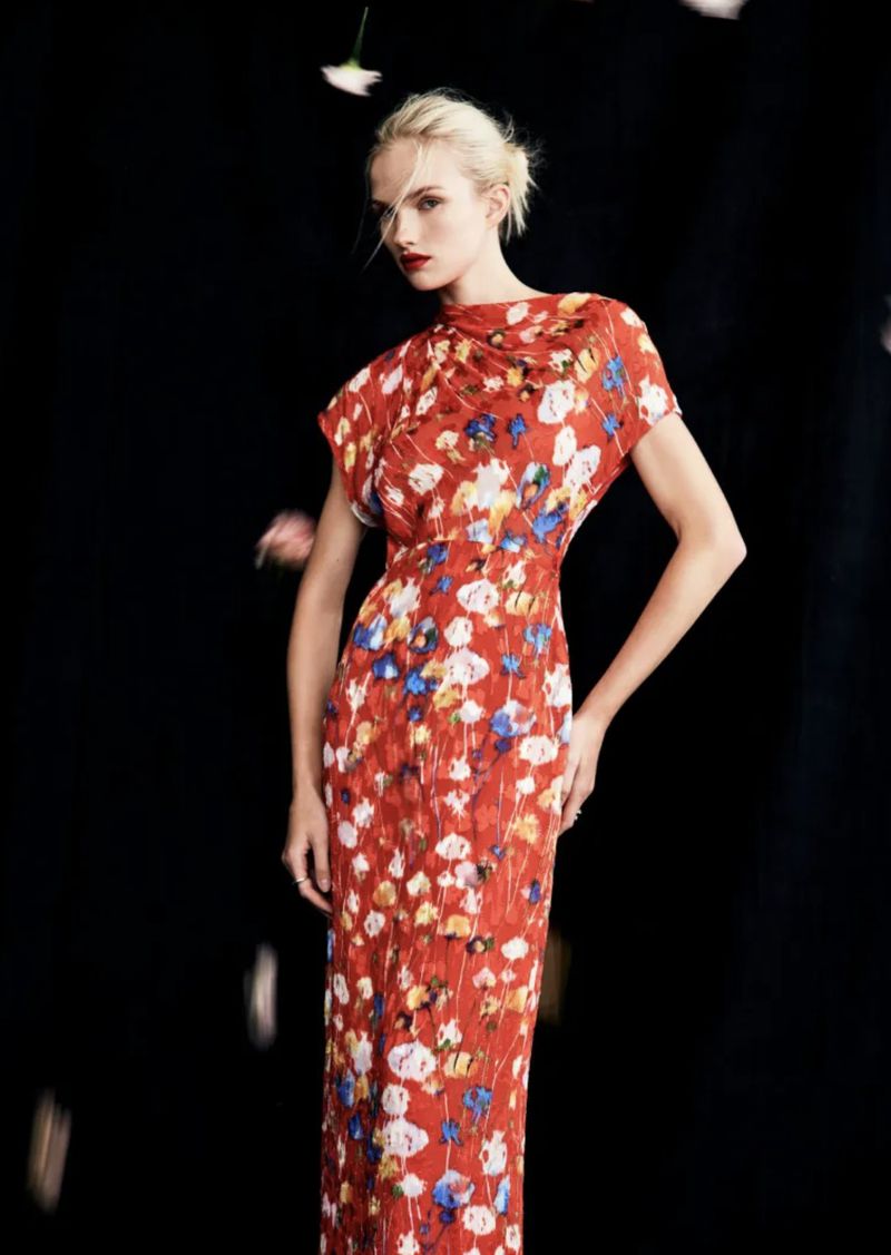 A woman models a floral-patterned gown with a black backdrop and falling flower petals, exuding high fashion and elegance.