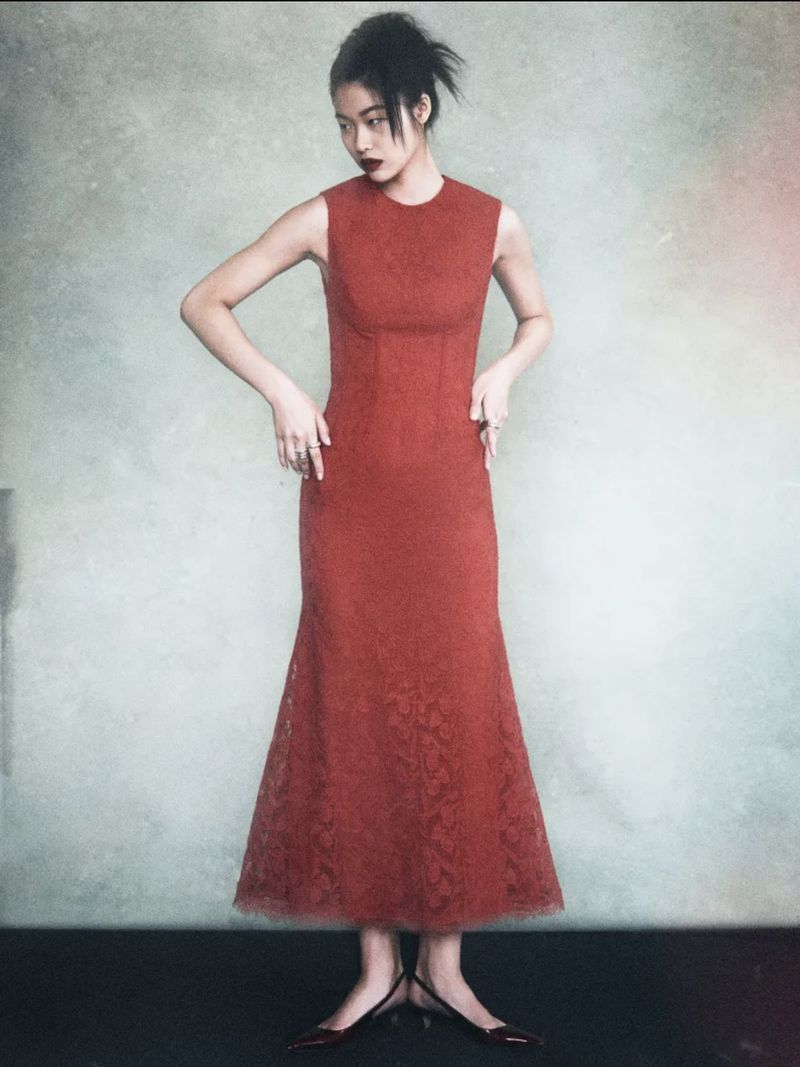 Full shot of an Asian woman in a sleeveless, rust-colored dress and heels standing in front of a mottled neutral background in a studio.