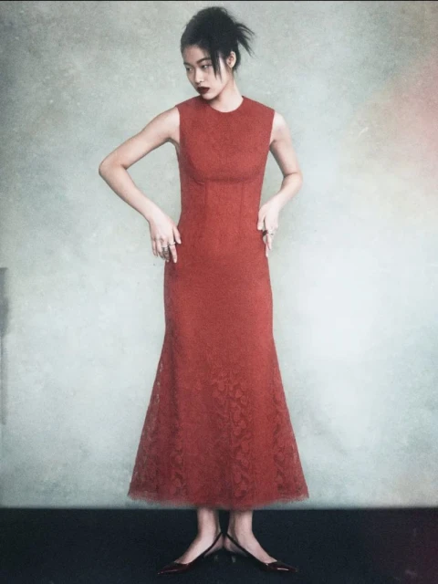 Full shot of an Asian woman in a sleeveless, rust-colored dress and heels standing in front of a mottled neutral background in a studio.