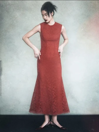 Full shot of an Asian woman in a sleeveless, rust-colored dress and heels standing in front of a mottled neutral background in a studio.