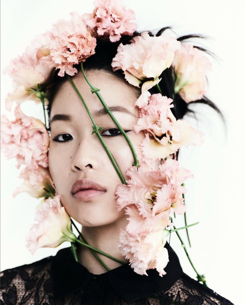 Portrait of an Asian woman with pink flowers in her hair, wearing a black lace top against a white background, in a beauty fashion setting.