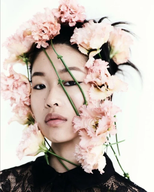 Portrait of an Asian woman with pink flowers in her hair, wearing a black lace top against a white background, in a beauty fashion setting.