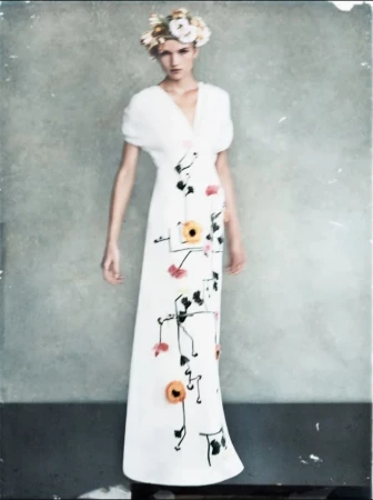 Model wearing a white floral dress with black botanical prints and a floral headpiece against a grey wall