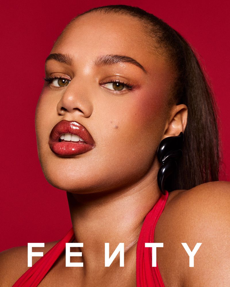 Close-up portrait of a woman with red makeup and glossy lips against a matching red background. She is wearing black earrings.