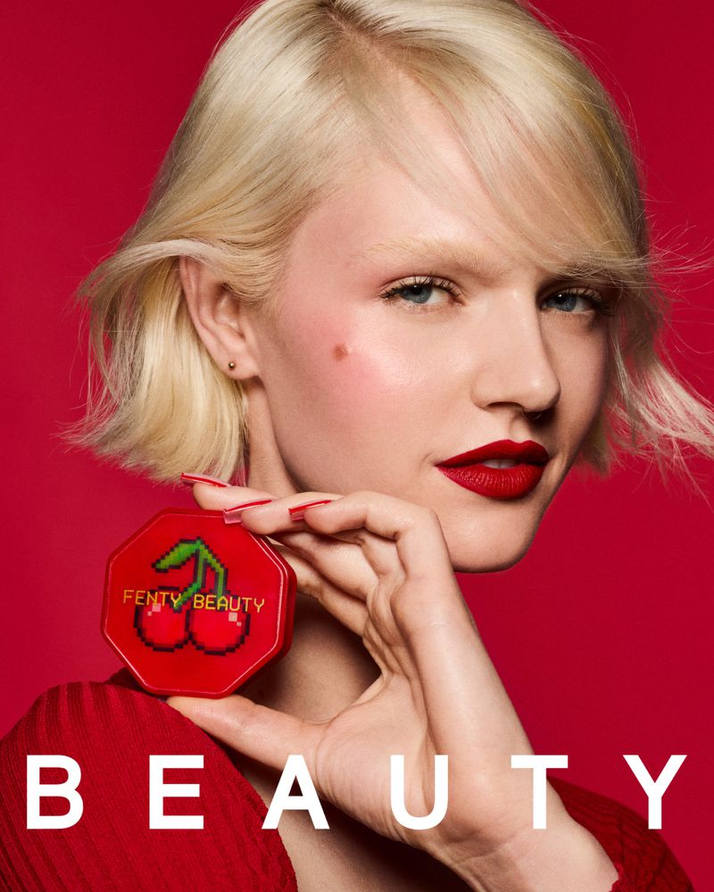 A fair-skinned adult female model with blonde hair is posing on a red background while holding a red Fenty Beauty makeup compact.