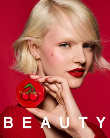 A fair-skinned adult female model with blonde hair is posing on a red background while holding a red Fenty Beauty makeup compact.