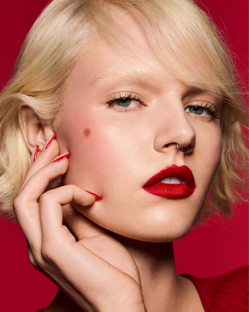 Close-up shot of a fair-skinned woman with blonde hair and red lipstick and nails, set against a red background in a beauty editorial.