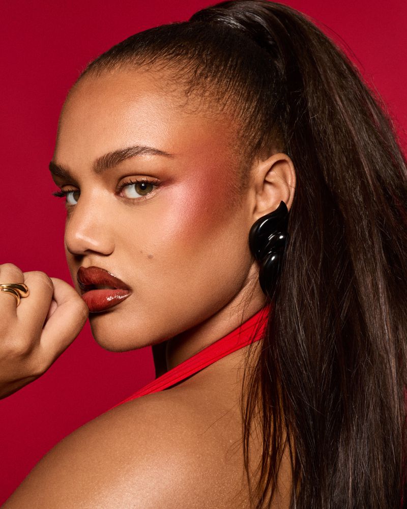 Close-up portrait of a young woman with dark hair in a high ponytail, wearing black statement earrings, and glossy red lipstick on a red background.