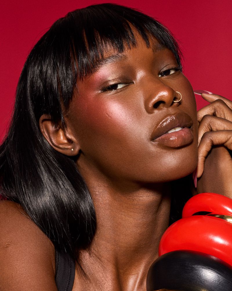 Close-up portrait of a dark-skinned woman with bangs and a nose ring wearing bracelets against a red background in a beauty shot.