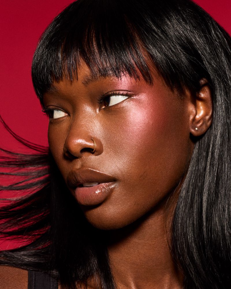 Close-up shot of a Black woman with glossy pink makeup, wearing a nose ring, and bangs, set against a red backdrop in a studio setting.