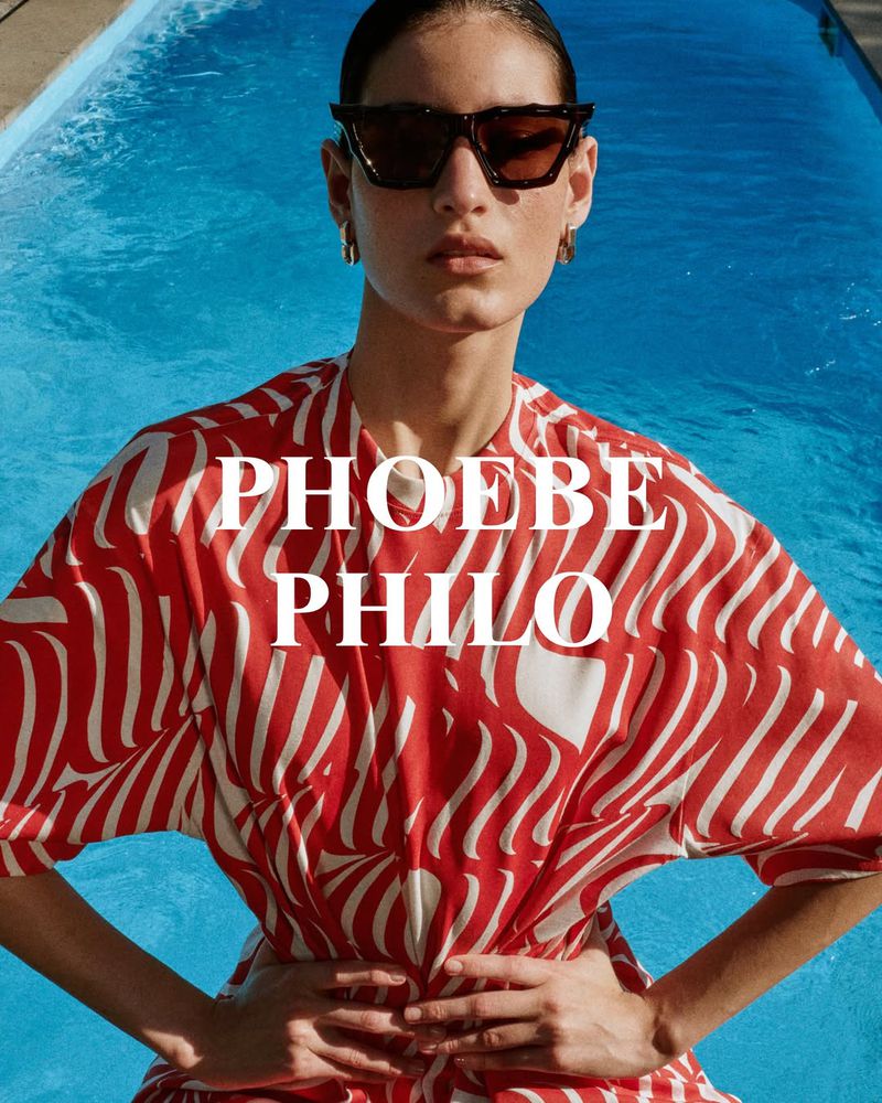 Woman with sunglasses poses in front of swimming pool wearing an orange and white patterned dress. Fashion shot near water.
