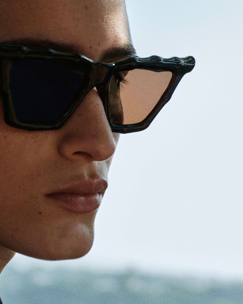 Close-up portrait of a model wearing dark sculptural sunglasses against an outdoor sky background
