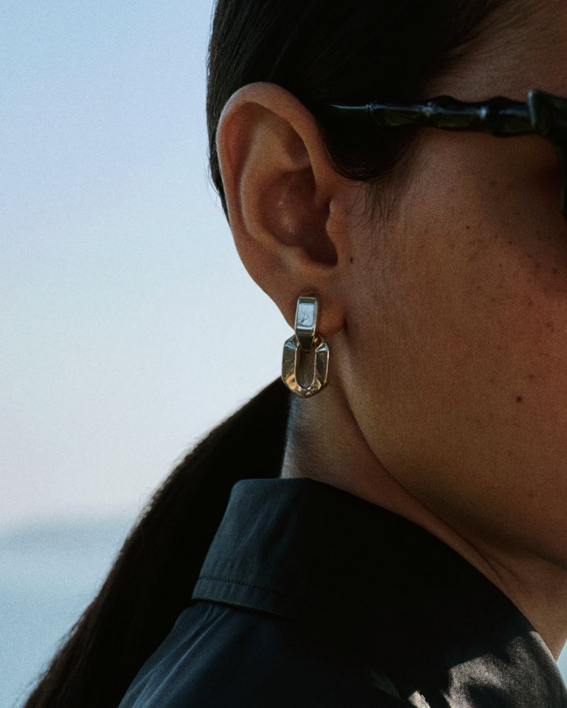 Close-up portrait of a model wearing metallic drop earrings and sunglasses against an outdoor sky background