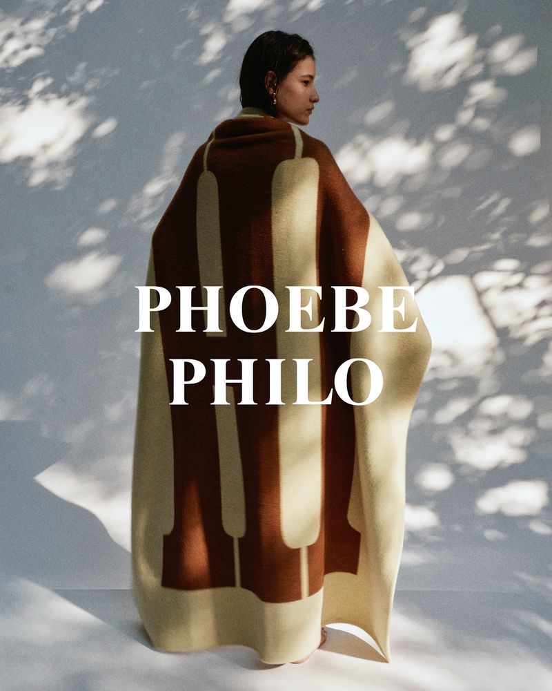 Woman with short brown hair draped in an off-white blanket with brown piano key design in a studio with shadow patterns.