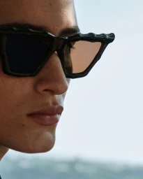 Close-up portrait of a model wearing dark sculptural sunglasses against an outdoor sky background
