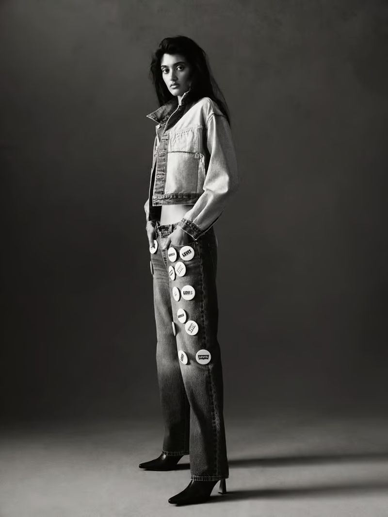 Studio shot of a young adult woman in a denim jacket and jeans decorated with circular badges, posing in front of a grey backdrop.