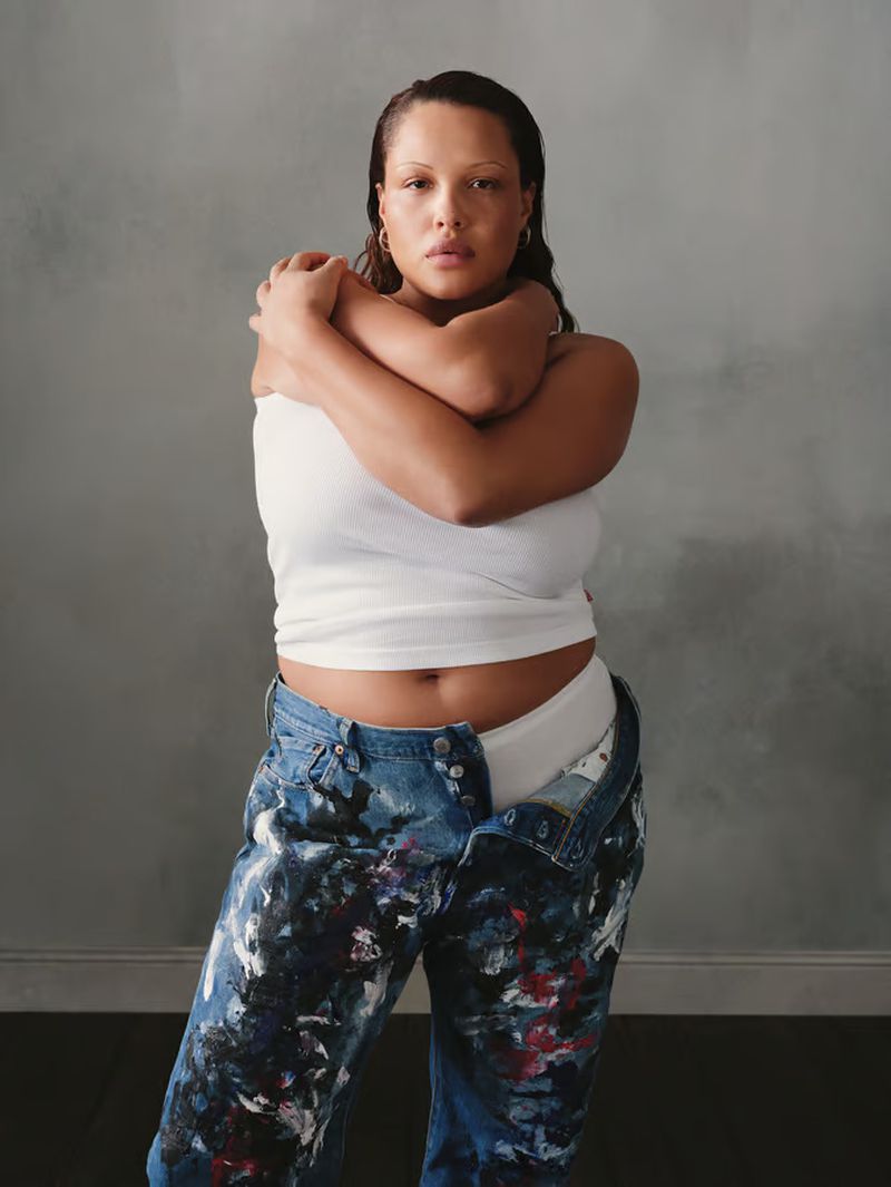 Woman with wet hair poses in a white tank top and paint-splattered jeans against a gray backdrop in a medium studio shot.