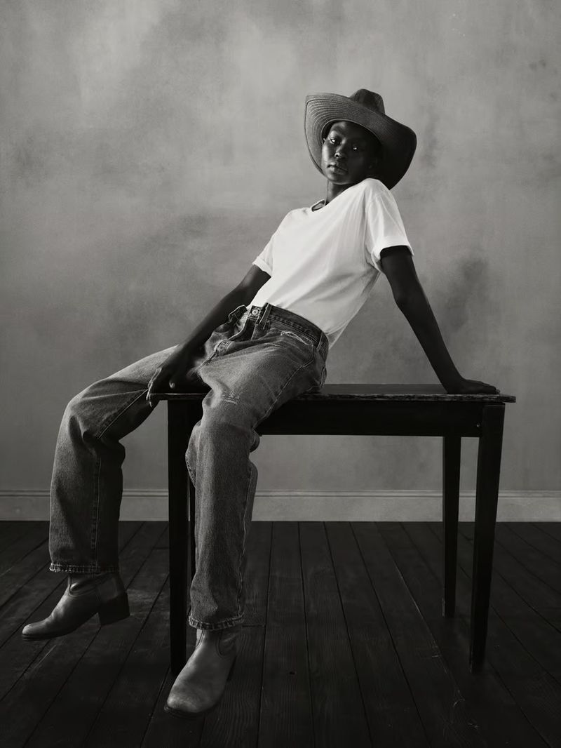Model wearing denim jeans, white t-shirt and cowboy hat sitting on a table in a studio portrait.