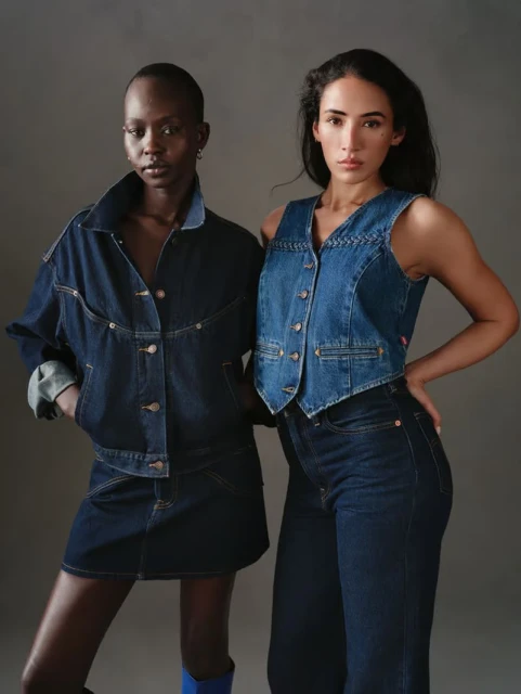 Two women pose in a photo studio wearing denim outfits. One is wearing a jacket and skirt, and the other is wearing jeans and a vest.