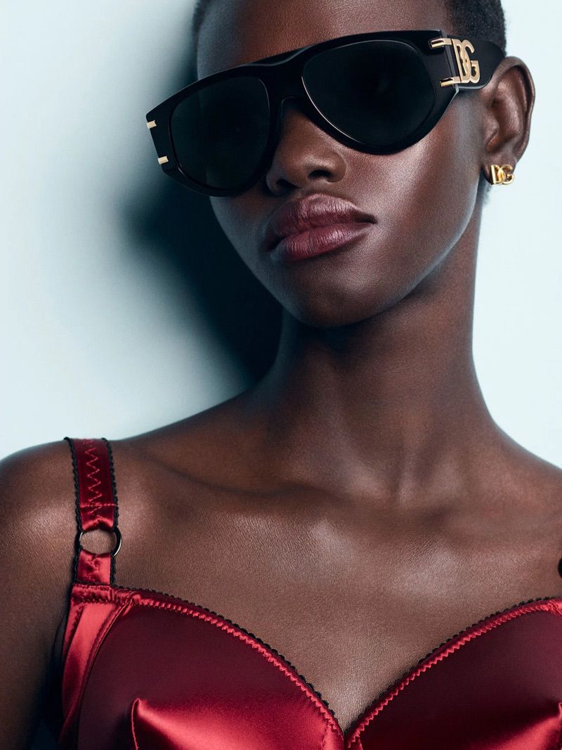 Close-up of an African-American woman wearing black sunglasses with DG logo and a burgundy satin bra, posing in a brightly lit studio.