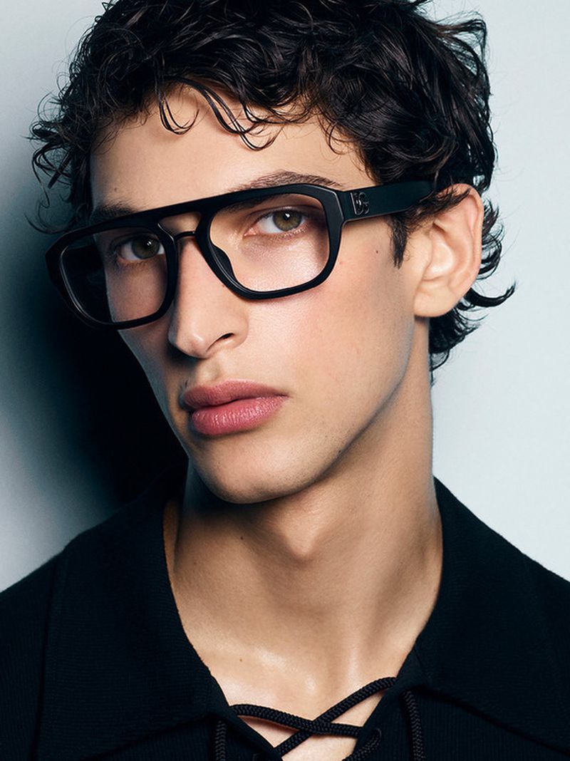 Close-up portrait of a young adult male with dark curly hair and wearing glasses, against a light blue background in a photo studio.