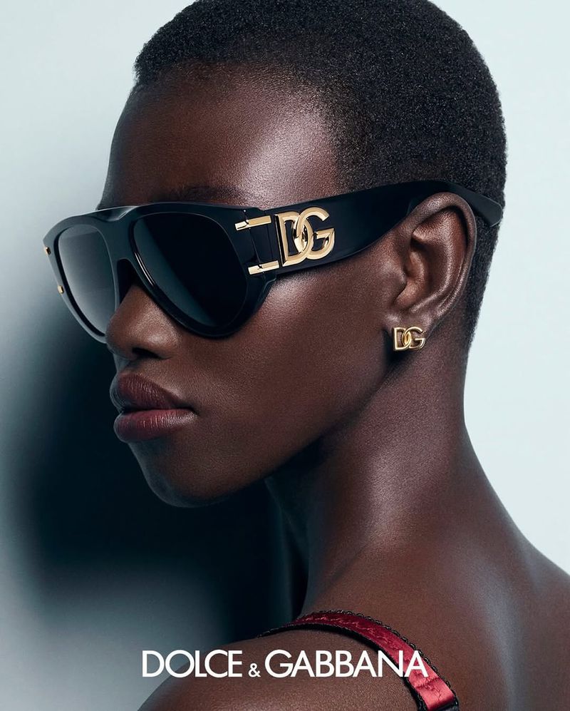 Close-up fashion portrait of a black woman wearing black sunglasses and gold earrings featuring the 'DG' logo, set against a light background.