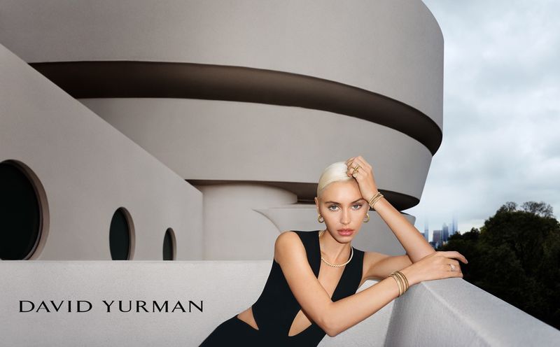Indoor shot of a blonde woman wearing a black dress and jewelry posed in front of a white circular building with a city skyline.