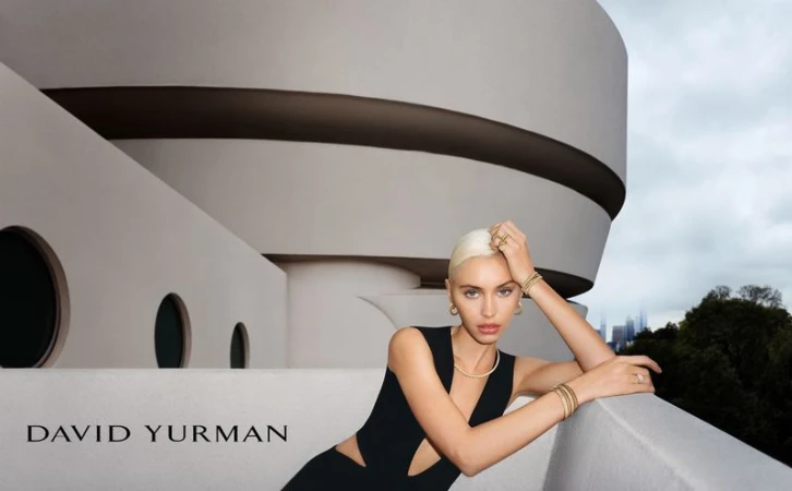 Indoor shot of a blonde woman wearing a black dress and jewelry posed in front of a white circular building with a city skyline.
