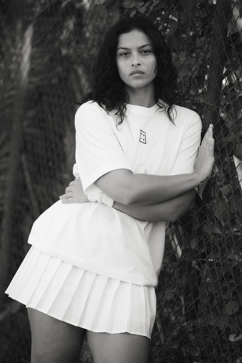 Monochrome portrait of a woman in a short white pleated skirt and t-shirt, leaning against a chain-link fence with foliage behind it.