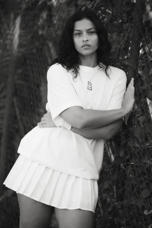 Monochrome portrait of a woman in a short white pleated skirt and t-shirt, leaning against a chain-link fence with foliage behind it.