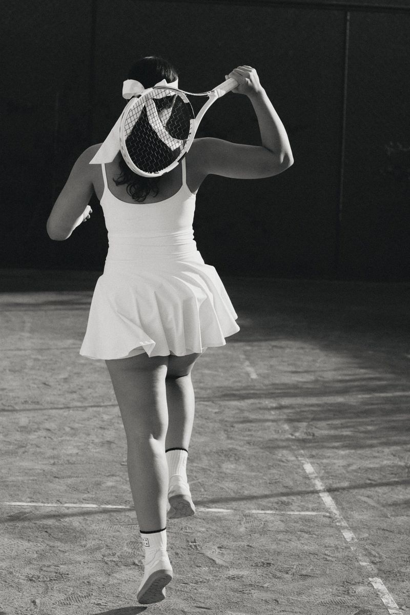 A woman in a white tennis outfit, viewed from behind, is playing tennis on a court, holding a tennis racket. Black and white photo.