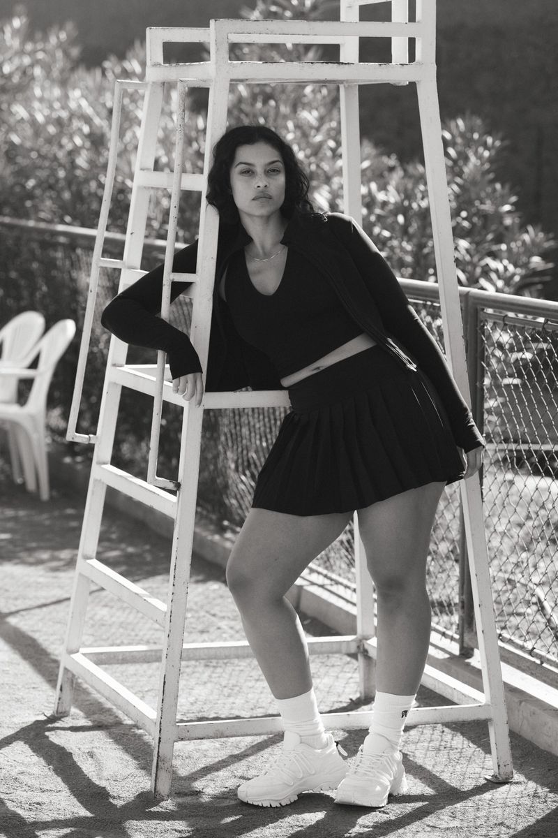 Woman in a black athletic outfit poses next to a white lifeguard chair on a tennis court outside; portrait-style black and white shot.