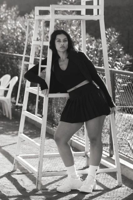 Woman in a black athletic outfit poses next to a white lifeguard chair on a tennis court outside; portrait-style black and white shot.