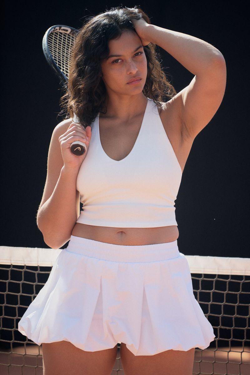 Portrait of woman in white tennis outfit holding a tennis racket on a tennis court with a dark background, natural lighting, and medium framing.