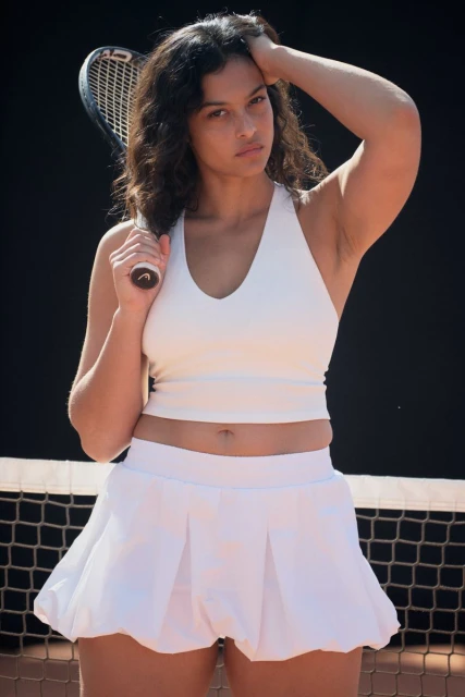 Portrait of woman in white tennis outfit holding a tennis racket on a tennis court with a dark background, natural lighting, and medium framing.