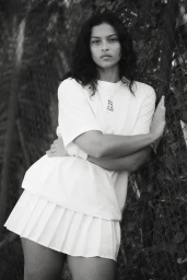 Model Devyn Garcia in a white t-shirt and pleated tennis skirt leaning against a chain-link fence outdoors in a black and white photograph