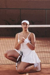 Model Devyn Garcia crouching on a clay tennis court holding a racket wearing a white tennis dress and visor