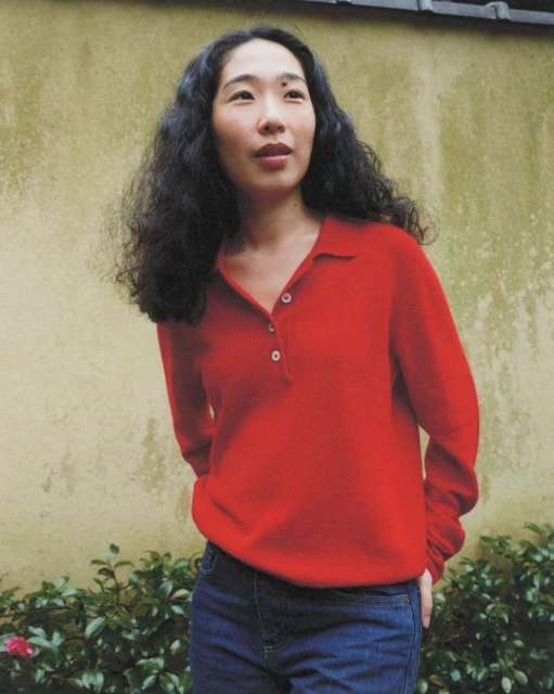 Woman in a red sweater and jeans stands outdoors in front of a weathered wall with foliage; medium shot, portrait orientation.