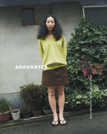 A woman in a yellow top and brown skirt stands outside with a building and greenery in the background, creating a casual fashion shot.