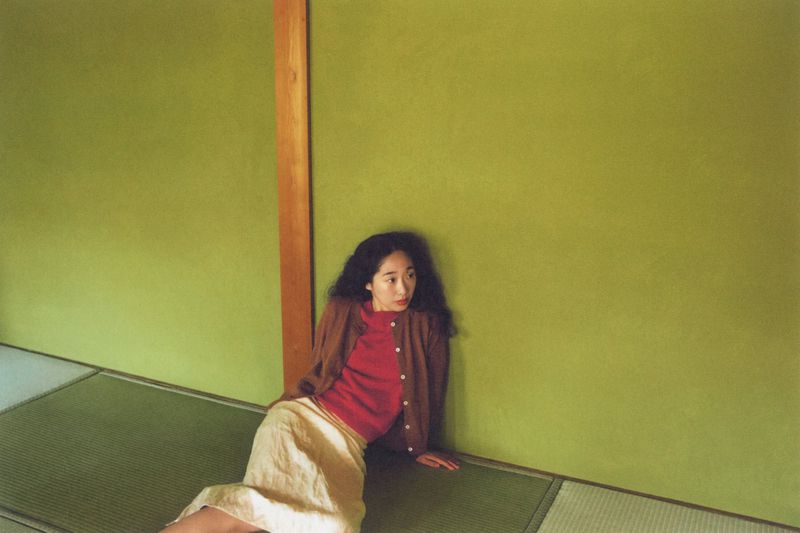 Young woman with dark curly hair and a red shirt sits on the floor against a green wall in a room with tatami mats.