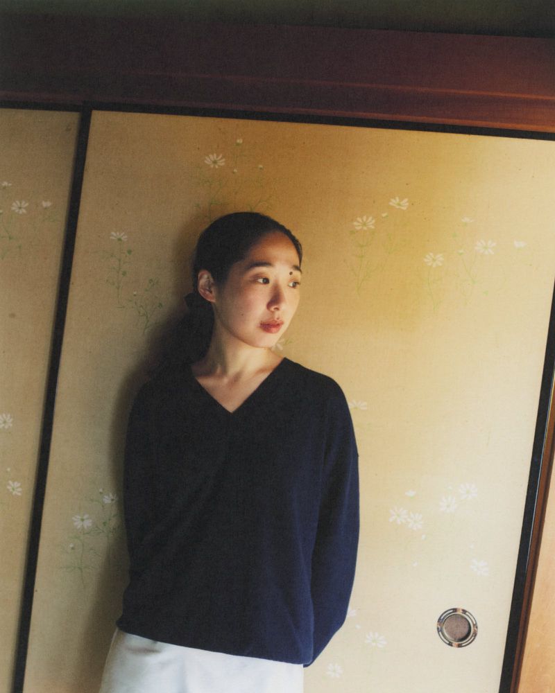 A young adult woman with dark hair stands indoors against a decorative screen, wearing a dark V-neck top and light skirt in soft lighting.