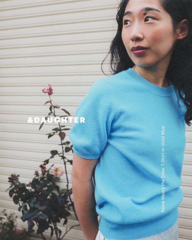 A stylish Asian woman in a bright blue short-sleeved sweater looks to the side; a lush rose bush and neutral siding complete the scene.