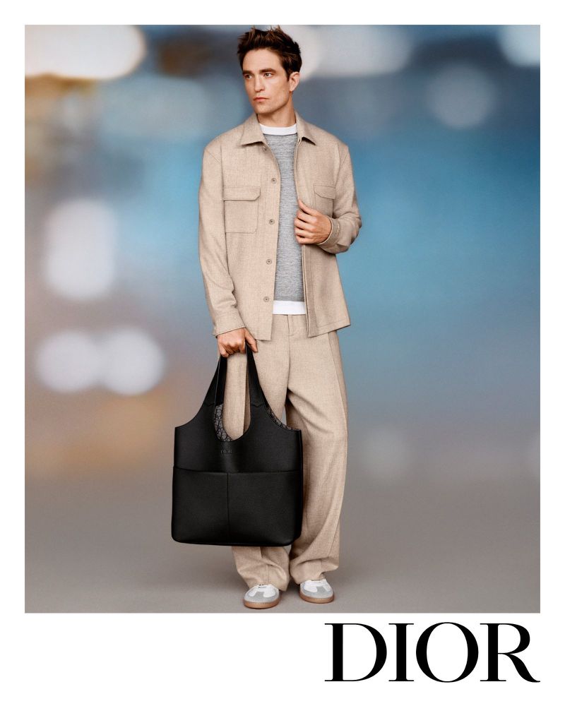 An adult man is shown wearing a tan outfit and holding a large black tote bag against a blurred background in a studio fashion shot.