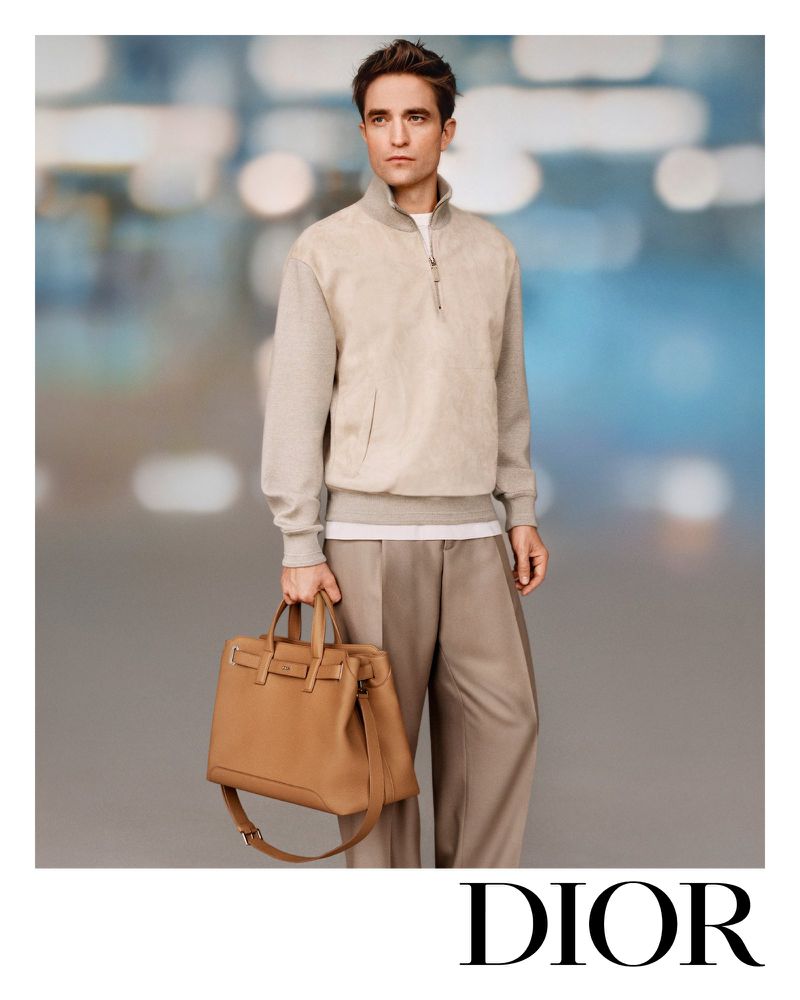 An adult male poses holding a caramel-colored leather tote bag. He is wearing beige trousers and a neutral-toned half-zip sweater.