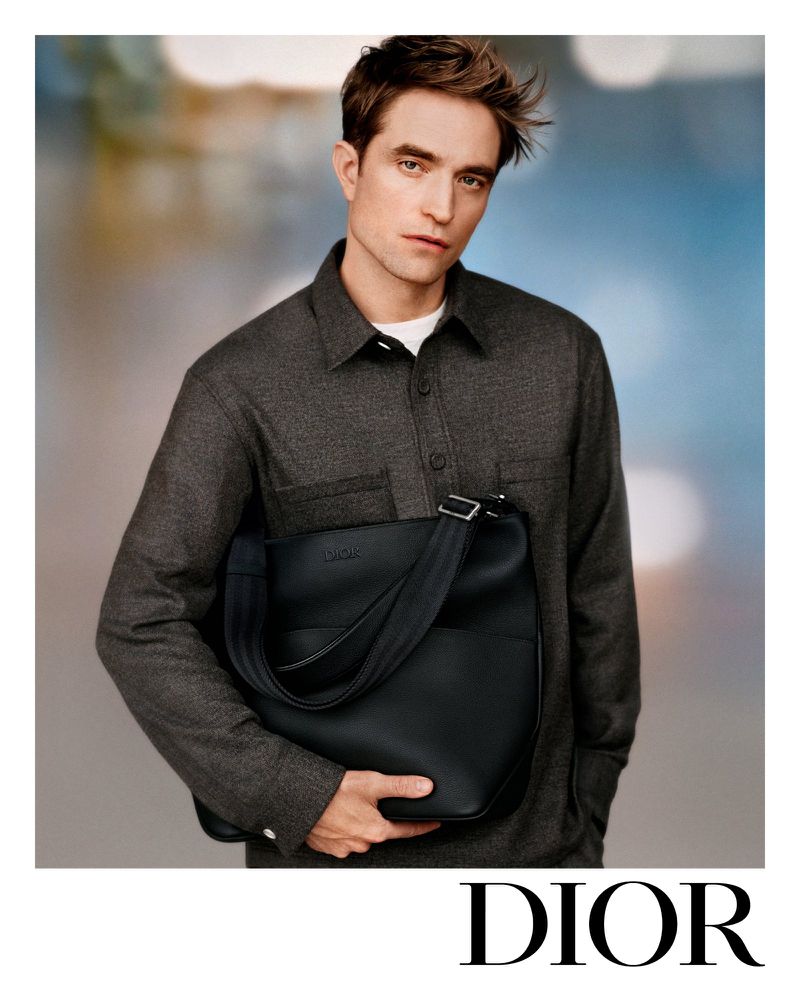 Man poses holding black leather Dior bag in a studio setting wearing a gray button-down shirt in a fashion forward portrait.